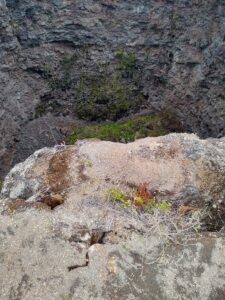 A view into the Devil's Throat pit crater. A rocky ledge dominates the foreground, with steep walls of rock in the background. A sliver of the crater bottom, covered in rubble, is visible