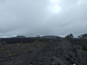 A large expanse of gray rock, with two peaks, one of which is Maunaulu, visible in the background. The sky is dark and cloudy.