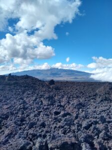 In the foreground, a field of rocky, gray, solidified lava sits. In the background, Mauna Kea sits in the midst of some clouds and the bright, blue sky.