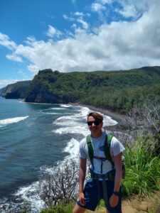 A young man standing in front of a coastline of green, lush valleys and white surf, with a clear, blue ocean to the left