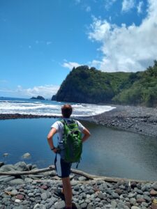 A young man wearing a green backpack, standing with his back to the camera. Beyond him is a clear, blue ocean on the left and a green, mountainous area on the right.