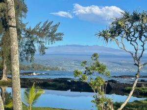 Mauna Kea, a massive volcanic mountain, looms in the background. Palm trees and the ocean are in the foreground.